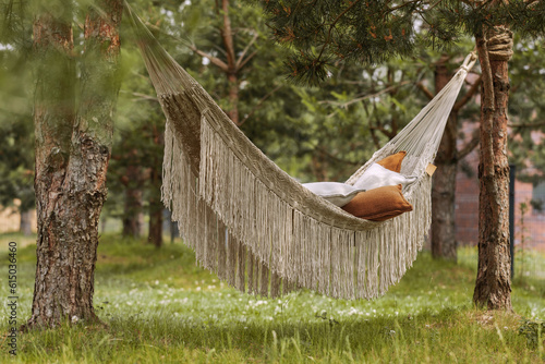 A neutral coloured woven hammock with fringes made from recycled plastic bottles hanging between two pines. The concept of  outdoor recreation, relaxation. Concept of recycle photo