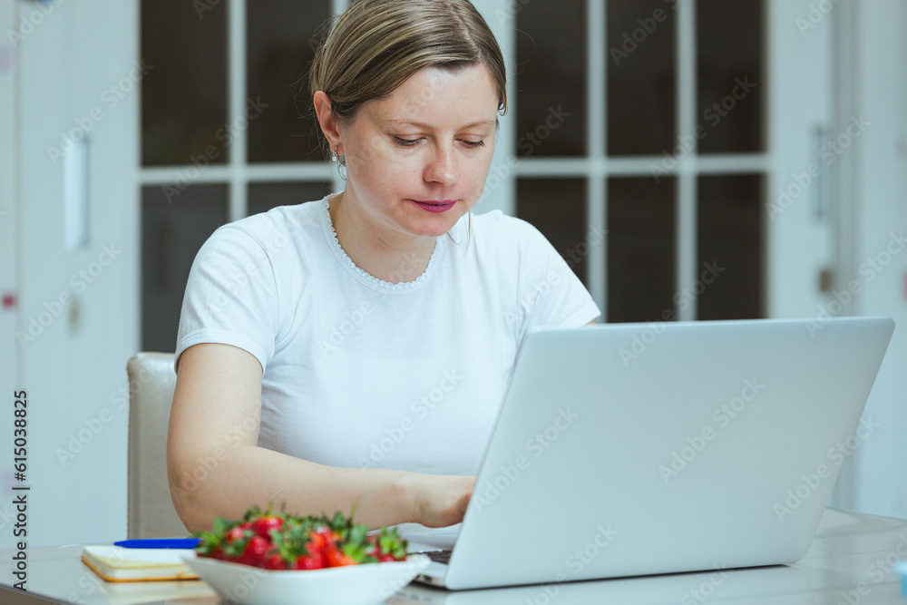 Woman working on a laptop and eating ripe strawberry for lunch, healthy eating seasonal fruits. Young woman studying online and having snack.
