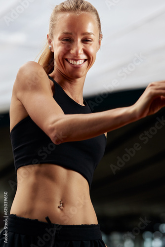 Fototapeta Naklejka Na Ścianę i Meble -  Young fitness girl is warming up in the stadium. Attractive slender smiling blonde in a black tracksuit makes active movements with her arms and legs. Active lifestyle.