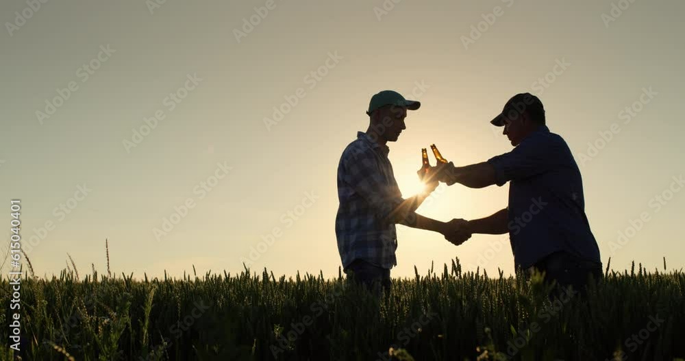 Silhouettes of two men clink bottles of beer and shake hands. Standing in a field of wheat at sunset Silhouettes of two men clink bottles of beer and shake hands. Standing in a field of wheat at sunset