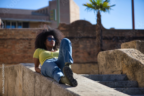 Young, beautiful, black woman with afro hair, wearing a yellow t-shirt, jeans and sunglasses, lying on a stone wall, relaxed, calm and sunbathing. Vacation concept, travel, current, modern.