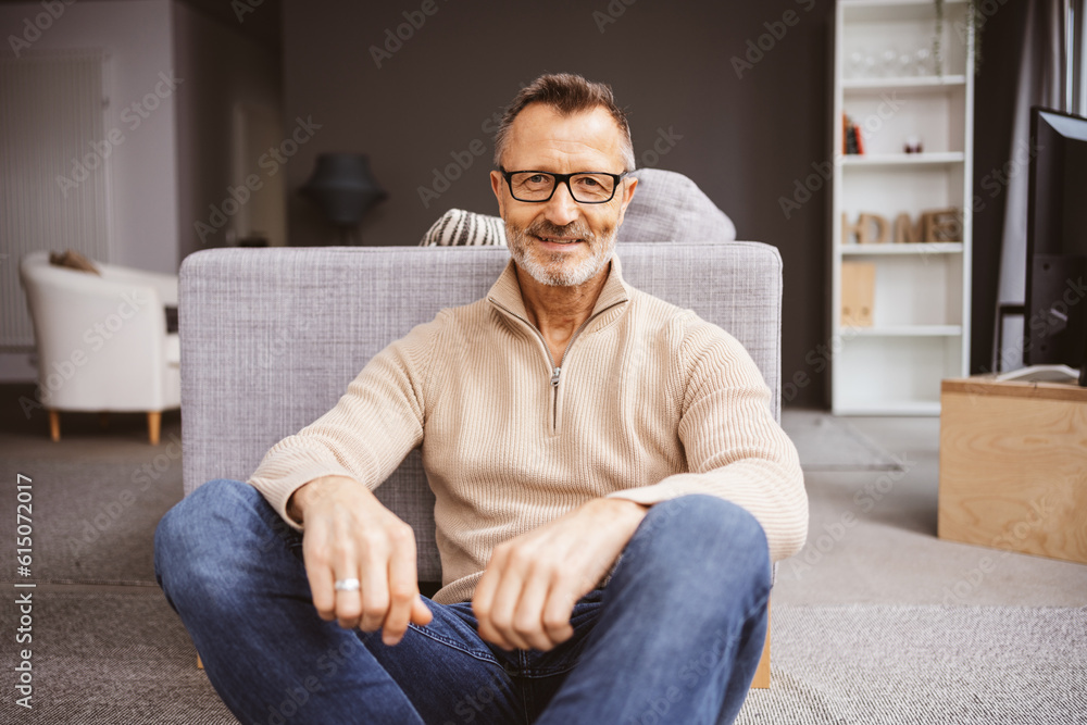 An older man sits on the floor in the living room and looks into the camera.