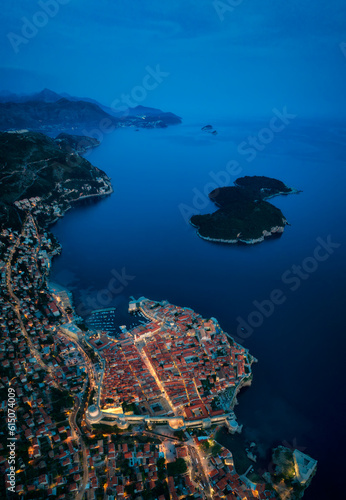 Amazing aerial panoramic view of the picturesque town of Dubrovnik with the old town, illuminated streets and buildings and marina with boats in blue hour after sunset.