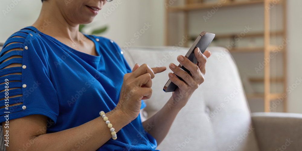 Smiling older asian woman sit rest on couch at home text or message online on modern cellphone gadget. Happy mature female relax browse internet on smartphone device. Elderly technology concept