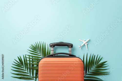 Summer escape calling! Top view shot of an orange suitcase, small airplane model, and palm leaves on a pastel blue background. Ample space available for text or advertising