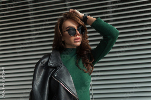 Stylish beautiful young lady with fashion sunglasses in trendy cool rock clothes with black leather jacket and green dress on the street near metal lines background