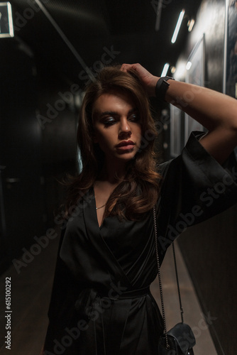 Fashionable beautiful chic woman in a stylish elegant black dress with bag in the dark indoors