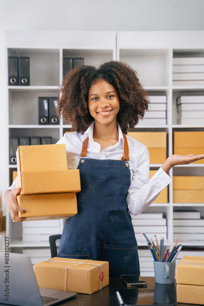 Young SMEs woman holding parcels and looking to camera, standing among several boxes in modern office. Young SMEs woman holding parcels and looking to camera, standing among several boxes in modern office.