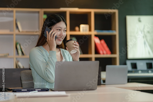 Attractive Asian woman talking on the phone with her business partner. Manage business to call marketing consultants and use laptop computers to analyze sales growth in the market.