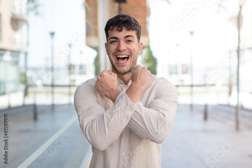 young handsome man smiling cheerfully and celebrating, with fists clenched and arms crossed, feeling happy and positive