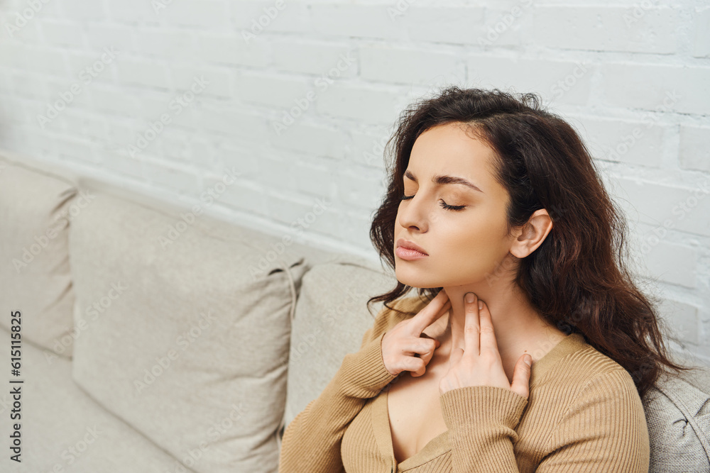 Young brunette woman with closed eyes in casual jumper massaging lymphatic nodes on neck during self-massage while sitting on couch at home, self-care ritual and holistic wellness practices concept Young brunette woman with closed eyes in casual jumper massaging lymphatic nodes on neck during self-massage while sitting on couch at home, self-care ritual and holistic wellness practices concept