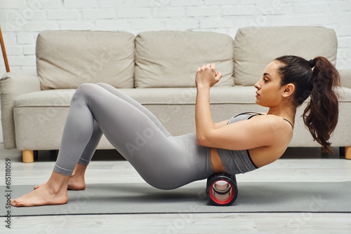 Side view of young and barefoot woman in sportswear massaging small of back with roller massager on fitness mat at home, sense of tranquility and promote relaxation concept, myofascial release photo