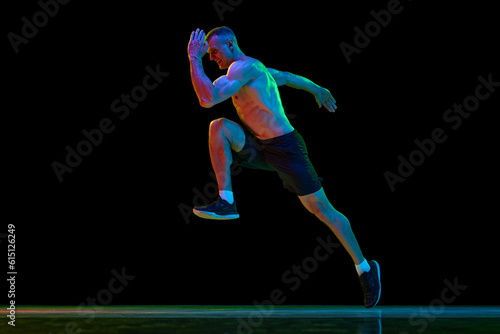 Young man with fit, muscular, relief body shape, professional sportsman in motion, running against black studio background in neon light