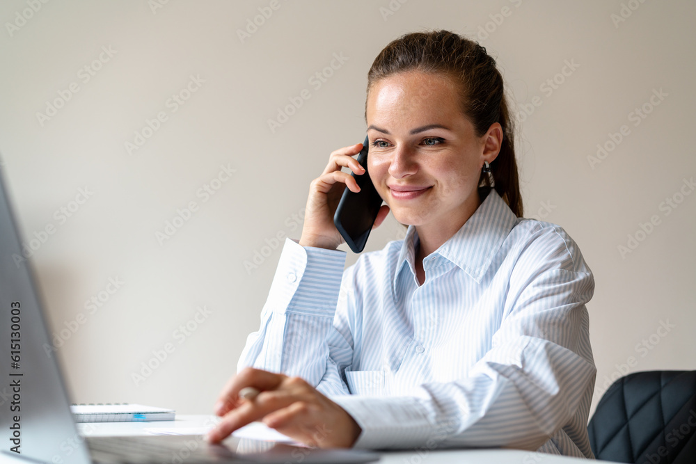 Woman white collar worker talking mobile phone and using her laptop while sitting at the desk in the office. Woman white collar worker talking mobile phone and using her laptop while sitting at the desk in the office.