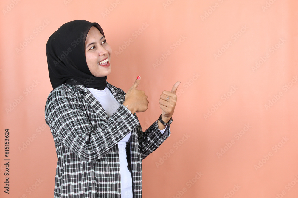Smiling beautiful Asian muslim woman pointing finger to empty space wearing a striped gray jacket and veil isolated peach background Smiling beautiful Asian muslim woman pointing finger to empty space wearing a striped gray jacket and veil isolated peach background