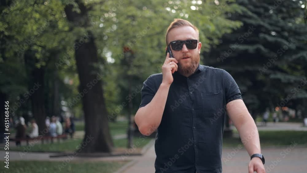 Close-up of young bearded Caucasian businessman in sunglasses walking in city and talking on mobile phone with business partner. Close-up of young bearded Caucasian businessman in sunglasses walking in city and talking on mobile phone with business partner.