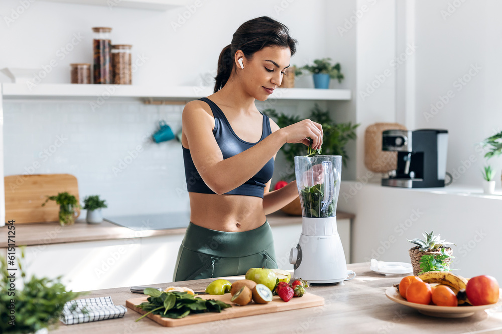 Athletic woman preparing smothie with vegetables and fruits while listening music with earphones in the kitchen at home.