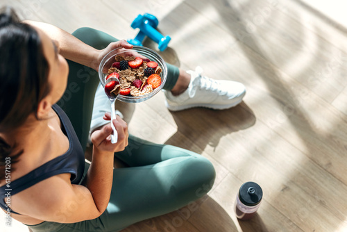 Athletic woman eating a healthy bowl of muesli with fruit sitting on floor in the kitchen at home photo
