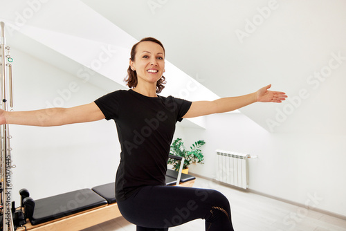 A woman is doing Pilates on a reformer bed in a bright studio. A slender flexible brunette in a black bodysuit is doing exercises to stretch the muscles. The concept of a healthy lifestyle.