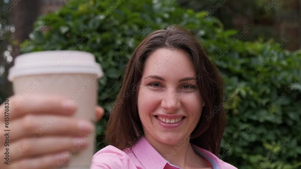 An attractive woman looks into the camera, smiles, picks up a paper cup and brings it to the camera against the background of a grass fence. Coffee advertising An attractive woman looks into the camera, smiles, picks up a paper cup and brings it to the camera against the background of a grass fence. Coffee advertising