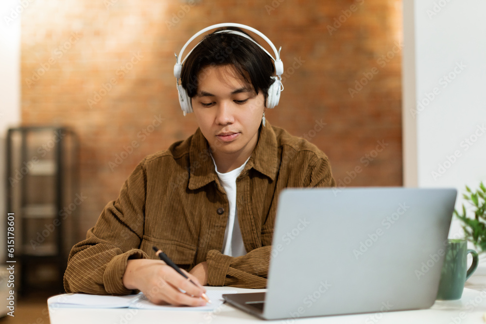 Asian Student Guy With Earphones Writing Notes Near Laptop Indoor Asian Student Guy With Earphones Writing Notes Near Laptop Indoor