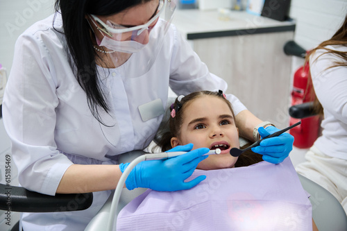 Female dentist doctor in medical protective eyewear  holding dental tools  performing dental treatment and cleaning to a little child girl in dentist s chair  curing caries in dentistry clinic