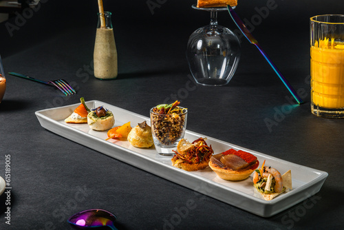 French sandwich placed on a returned wine glass,  various gastronomic appetizers placed on a white  rectangular plate , glass of orange juice, table cutlerys, original setup on a black background photo