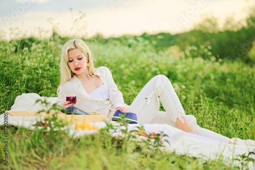 A blonde woman with a glass of wine lounges on a picnic. Lady lying on a blanket on the grass © Olek