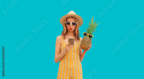 Summer portrait of stylish happy young woman with smartphone and fresh pineapple wearing straw hat, sunglasses on blue background