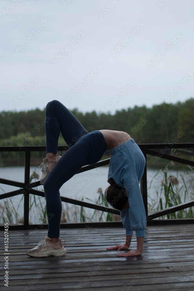 View of a girl with athletic body in blue gym suit is doing acrobatics exercises near lake.