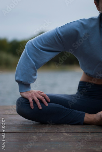 View of a girl with athletic body in blue gym suit is doing yoga exercises in cloudy rainy weather near lake.