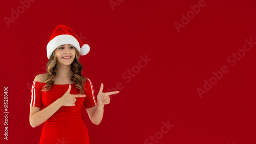 Smiling young Santa woman in red dress, Christmas hat pointing index fingers aside on mock up copy space isolated on red background, studio portrait. Happy New Year celebration merry holiday concept