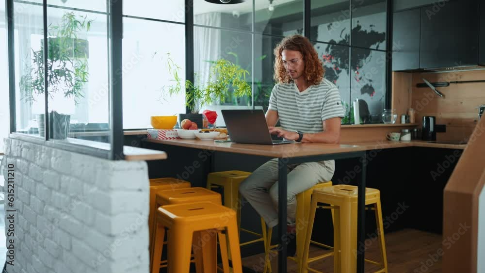 Smiling curly haired man in eyeglasses working on laptop in office kitchen Smiling curly haired man in eyeglasses working on laptop in office kitchen