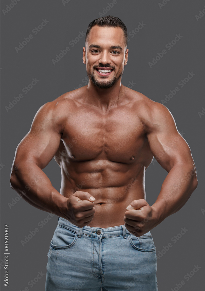 Fototapeta premium Incredibly attractive male model with a well-toned physique flashes a bright smile as he poses shirtless against a simple grey background, exuding charisma and magnetism