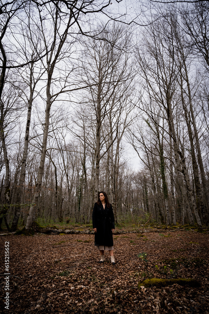 A young woman alone walking through a beech forest with a blanket of leaves on the ground dressed in a long black coat A young woman alone walking through a beech forest with a blanket of leaves on the ground dressed in a long black coat