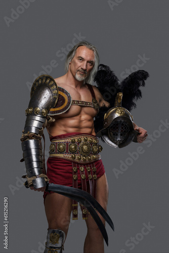 Imposing gladiator of mature age with a stylish silver beard and flowing grey hair dons lightweight armor, wielding a gladius and holds helmet as he stands proud against a neutral grey backdrop