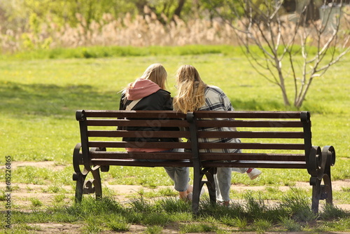 Two girls are sitting on a bench in the park in the summer. Outdoor recreation far from the bustle of the city. Stylish women chatting in a relaxed environment among green spaces