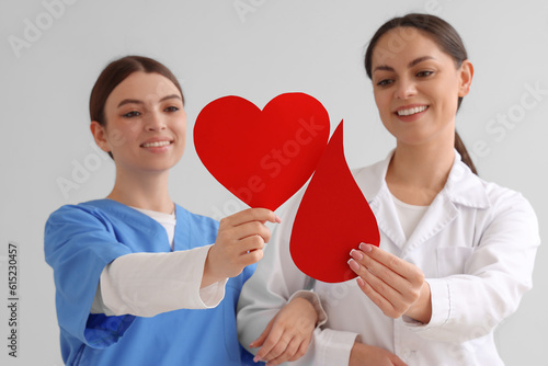 Female doctor with nurse holding paper heart and blood drop on light backgrou...