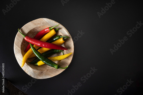 Multicolored pods of hot chili peppers lie on a wooden textured plate tray bowl on a black table