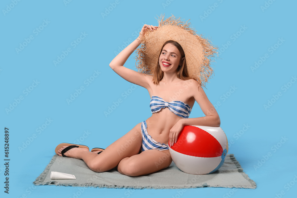 Young woman in swimsuit with beach ball sitting on blue background