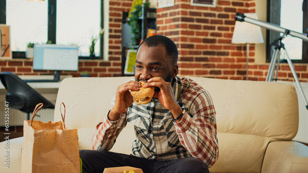 Modern adult having hamburger and fries for dinner, eating fast food meal from takeaway and watching movie on television. Young guy enjoying burger and drinking bottle of beer at home.