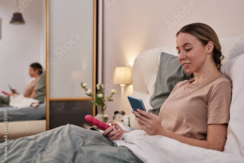 Side view portrait of smiling young woman holding sex toy and smartphone while sitting in bed at home, copy space