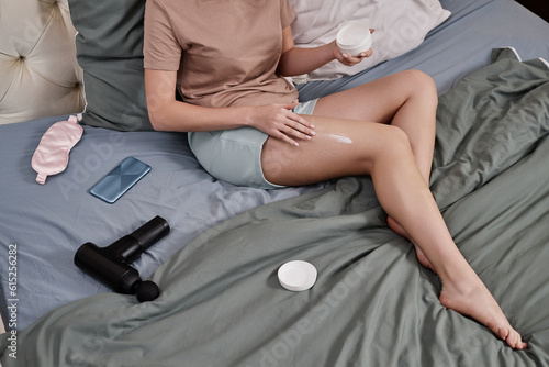 Unrecognizable young woman enjoying self care day at home and using body cream while sitting on bed, copy space