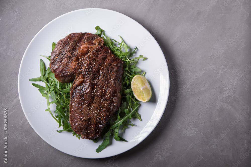 Ribeye steak with arugula on a white plate with lime on a grey background, top view.