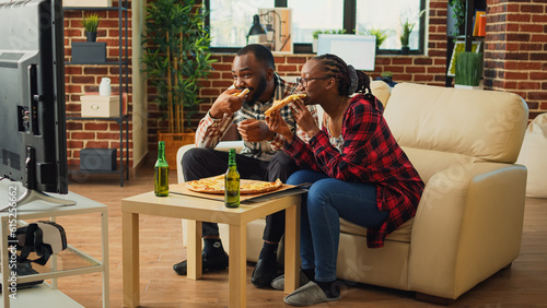 Bild auf Leinwand Happy partners eating slices of pizza on sofa and binge watching tv show at home, enjoying fast food meal