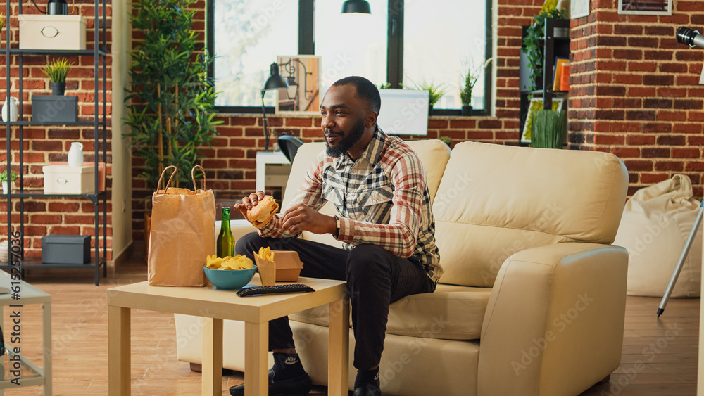 Cheerful man taking bite of hamburger in living room, eating fast food from takeaway delivery order. Happy adult watching television at home and serving burger with fries and alcohol. Cheerful man taking bite of hamburger in living room, eating fast food from takeaway delivery order. Happy adult watching television at home and serving burger with fries and alcohol.