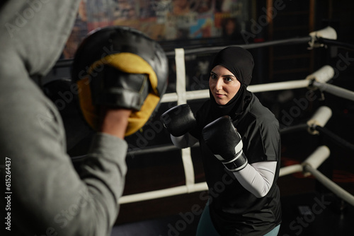 Young Muslim woman in hijab, activewear and leather boxing gloves looking at her trainer with defense pads on hands during training
