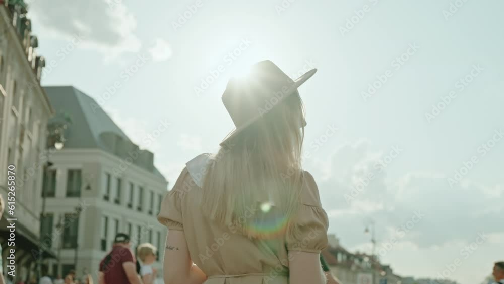 Happy relaxed female wearing beige dress and hat walking outdoors in old town street enjoys sunny weather, following slow motion young blonde hair woman turns around backlit bright evening sunlight Happy relaxed female wearing beige dress and hat walking outdoors in old town street enjoys sunny weather, following slow motion young blonde hair woman turns around backlit bright evening sunlight