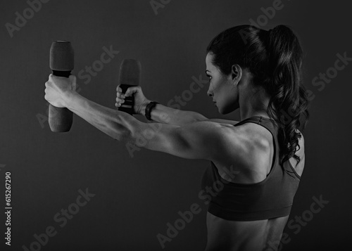 Female sporty muscular young serious woman doing strength workout on the shoulders, biceps and arms in sport bra holding dumbbells on black background. Closeup back view.