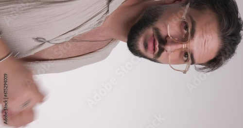 unshaved man with glasses looking down, adjusting necklace and moving in a side view pose in front of light grey background